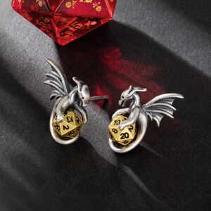 May include: A pair of silver dragon earrings with gold 20-sided dice. Each dragon is holding the die in its claws.