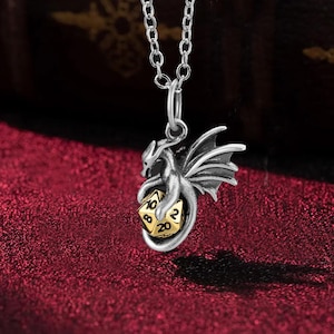 Puede incluir: Colgante de plata con un dragón que sostiene un dado dorado de veinte caras, suspendido de una cadena de plata. El dragón tiene alas detalladas y está sobre un fondo rojo oscuro. El dado muestra el número 20.