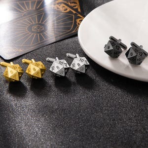 May include: A set of six cufflinks featuring a dice design. Three cufflinks are gold and three are silver. Each cufflink has a different number on each side. The cufflinks are arranged on a black surface.
