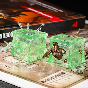 May include: Two green, cube-shaped resin miniatures with embedded figures and swords, designed for tabletop gaming. One cube features a warrior figure, while the other has multiple swords protruding from the top. The miniatures are set on a game board.