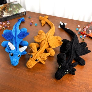 Puede incluir: Tres dragones de ganchillo hechos a mano en azul, dorado y negro. Cada dragón tiene ojos grandes, alas y una cola larga. El dragón azul tiene cuernos blancos. Los dragones están sobre una superficie de madera.