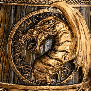 Op de afbeelding: Een close-up van een goudkleurig drakenreli&euml;f op een donkerbruine achtergrond. De draak staat in een cirkelvormig frame met sierlijke details. De draak heeft open kaken en grote vleugels, waardoor hij een woeste uitstraling heeft. Het item lijkt een decoratieve mok te zijn.