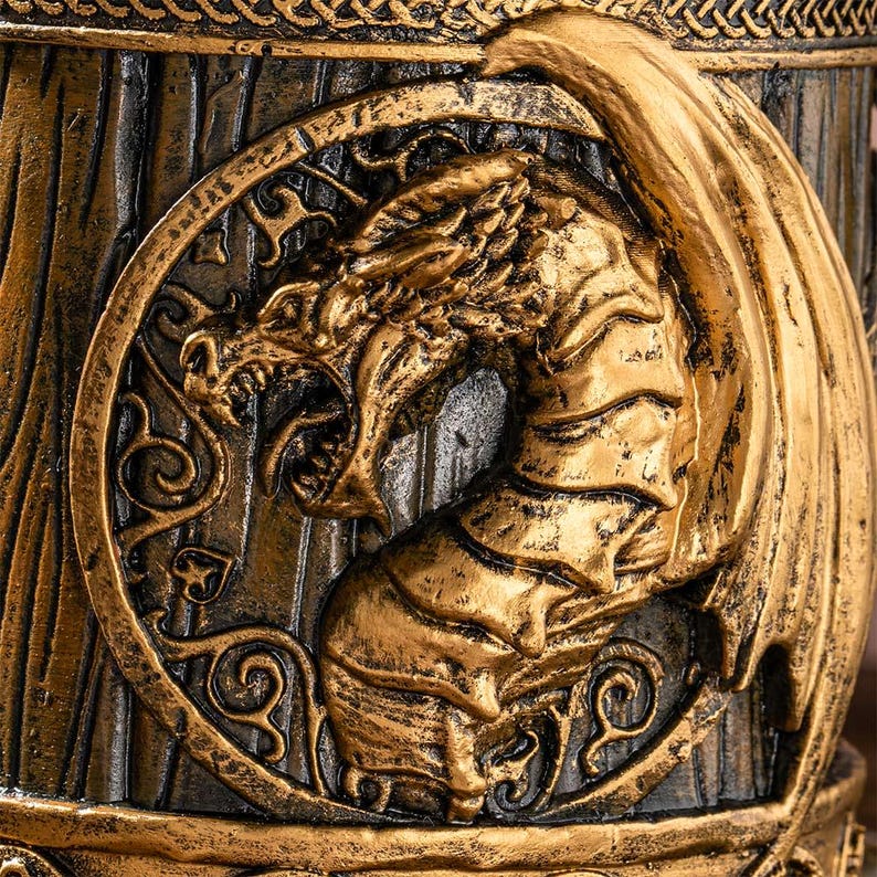 Op de afbeelding: Een close-up van een goudkleurig drakenreli&euml;f op een donkerbruine achtergrond. De draak staat in een cirkelvormig frame met sierlijke details. De draak heeft open kaken en grote vleugels, waardoor hij een woeste uitstraling heeft. Het item lijkt een decoratieve mok te zijn.