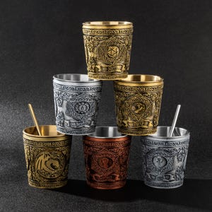 May include: A collection of six ornate metal shot glasses in various colors, including gold, silver, and copper. Each glass features intricate embossed designs and a unique emblem. Some glasses include small metal stir sticks.