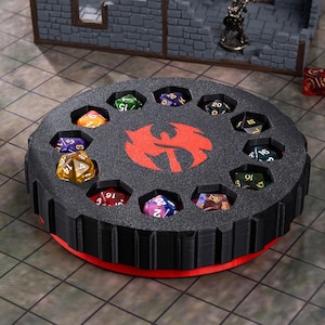 May include: A black dice storage container with a red logo and a red base. The container holds twelve multi-sided dice in individual compartments. The dice are various colors and show different numbers. The container is on a patterned surface.
