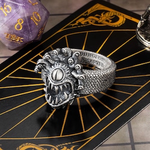 May include: A silver-colored ring featuring a detailed monster design with multiple eyes, sharp teeth, and the text "DUNGEON MASTER". The ring is set against a black background with gold lines, with a purple dice in the background.