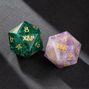 Puede incluir: Dos dados de piedra preciosa, uno verde y otro morado, con números dorados y el texto "X&P" en cada uno.