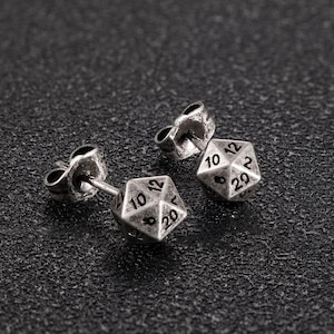 May include: Silver-coloured stud earrings in the shape of twenty-sided dice, with numbers 2, 8, 10, 12, and 20 visible. The earrings are designed for pierced ears and are shown on a textured, dark surface.