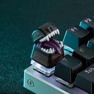 Puede incluir: Una tecla personalizada con forma de cofre del tesoro con boca de monstruo. La tecla es marrón con dientes blancos y una lengua morada. Está instalada en un teclado mecánico con teclas negras.