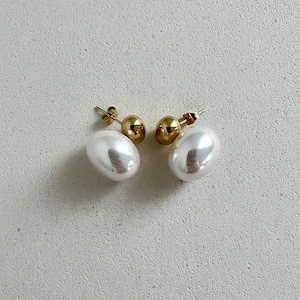 Peut inclure: Une paire de boucles d'oreilles en or et en perles blanches. Chaque boucle d'oreille présente une grosse perle blanche ronde avec une petite boule dorée ronde à l'arrière.
