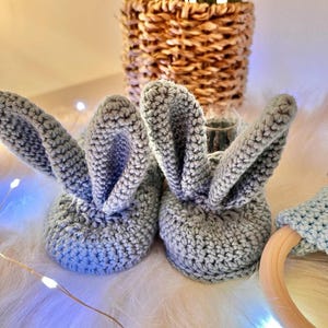 May include: A pair of light gray crocheted baby booties with bunny ears. The booties are made of soft yarn and are designed to keep a baby's feet warm. The ears stand up, adding a playful touch to the design.