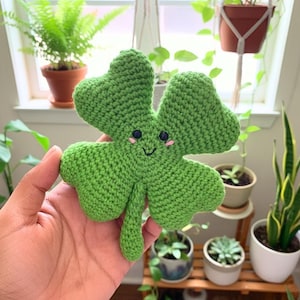 May include: A handmade, green crocheted four-leaf clover with a smiling face and pink cheeks. The clover is held in a hand, with a background of potted plants. This whimsical item is perfect for St. Patrick's Day.