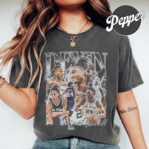 Pode incluir: Uma t-shirt de manga curta cinzento-carvão com uma impressão gráfica do jogador de basquetebol Devin Vassell em várias poses de ação. O design inclui o texto "DEVIN" no topo e "Vassell" na parte inferior, com estrelas e estilo vintage.