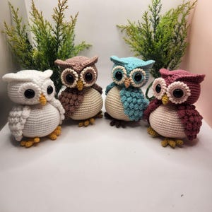 May include: Four crocheted owl figurines arranged in a row. Each owl features a round cream-colored belly, large black plastic eyes, a small yellow beak, and textured wings in white, brown, light blue, and dusty rose colors with yellow feet.