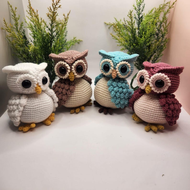 May include: Four crocheted owl figurines arranged in a row. Each owl features a round cream-colored belly, large black plastic eyes, a small yellow beak, and textured wings in white, brown, light blue, and dusty rose colors with yellow feet.