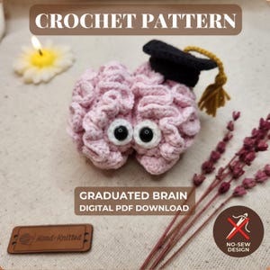 Op de afbeelding: Een lichtroze gehaakt hersenspeeltje met zwarte veiligheidsoogjes en een zwarte afstudeerhoed met een gouden kwastje. De afbeelding bevat de tekst CROCHET PATTERN, GRADUATED BRAIN DIGITAL PDF DOWNLOAD en een icoon voor naadloos ontwerp.