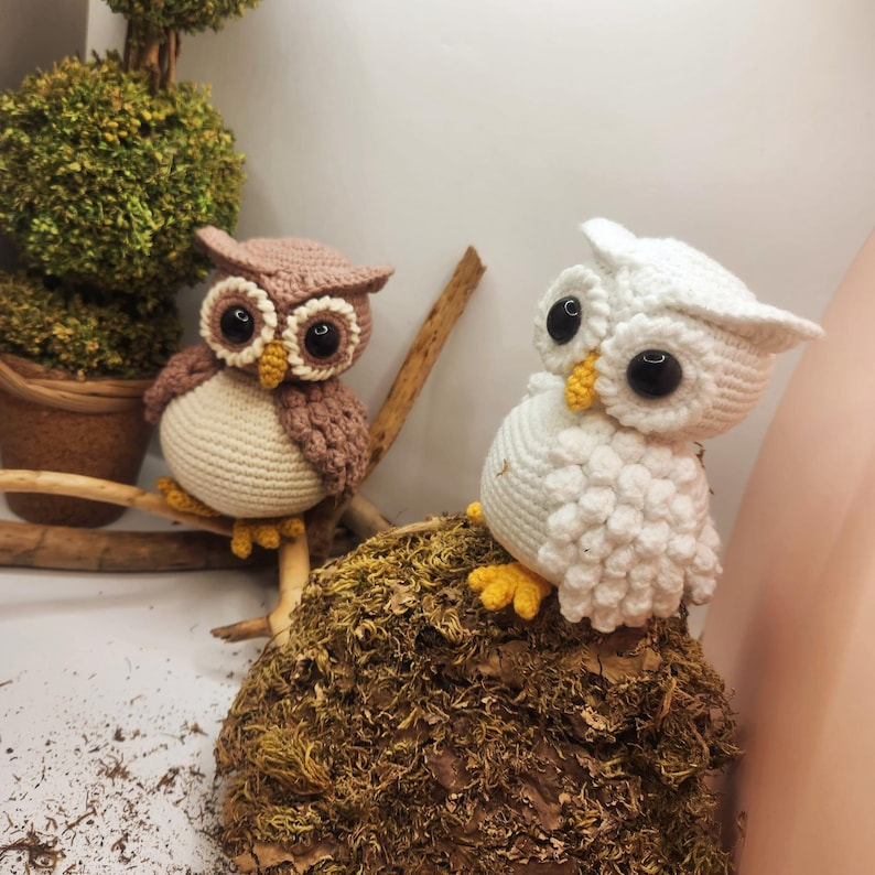 May include: Two crocheted owl figurines, one brown and cream and one white, each featuring large black plastic eyes, yellow beaks, and yellow feet. The brown owl sits on a wooden branch, while the white owl rests on a textured mossy surface.