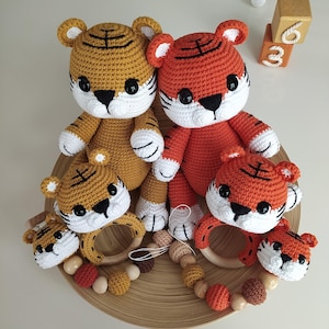 Puede incluir: Un conjunto de cuatro juguetes de tigre de crochet, dos naranja y dos marrones, con rayas negras. Los tigres sostienen anillos de dentición de madera. Un collar de cuentas de madera con cuentas naranja, marrón y de madera está en primer plano.