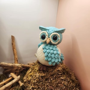 May include: A light blue and cream-colored crocheted owl figurine featuring large black plastic eyes, a small tan beak, and textured wing details. The owl has a round body with a cream-colored belly and light blue head and wings.