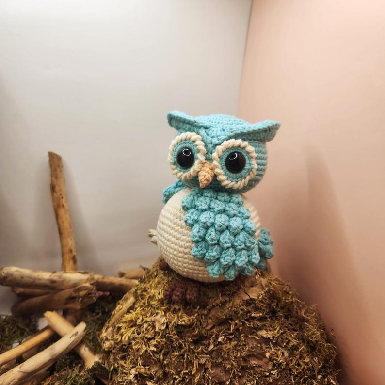 May include: A light blue and cream-colored crocheted owl figurine featuring large black plastic eyes, a small tan beak, and textured wing details. The owl has a round body with a cream-colored belly and light blue head and wings.