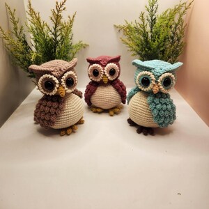 May include: Three crocheted owl figurines featuring large black eyes, yellow beaks, and cream-colored bellies. The owls are colored in shades of brown, dusty rose, and light blue, each with textured wing details and small feet.