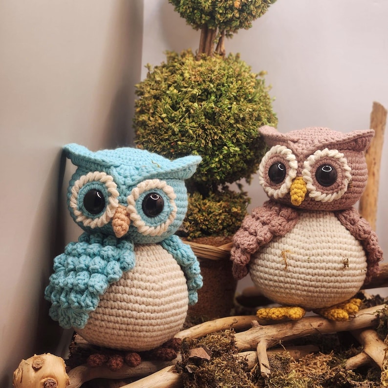 May include: Two crocheted owl figurines. One owl is light blue with a cream-colored belly, while the other is dusty rose with a cream-colored belly. Both feature large black plastic eyes, textured wing details, and small yellow or tan beaks.