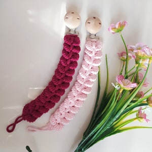 May include: Two handmade pacifier clips. One is a deep burgundy color, and the other is a soft pink. Both have a wooden clip and a crocheted design. The clips are next to a bouquet of pink flowers and green leaves.