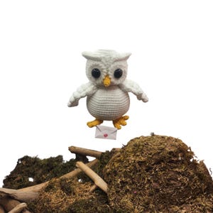 May include: A white crocheted owl plush toy with large black eyes, a yellow beak, and yellow feet. The owl is holding a small white envelope with a red wax seal in its talons. The texture of the yarn is visible throughout the body.