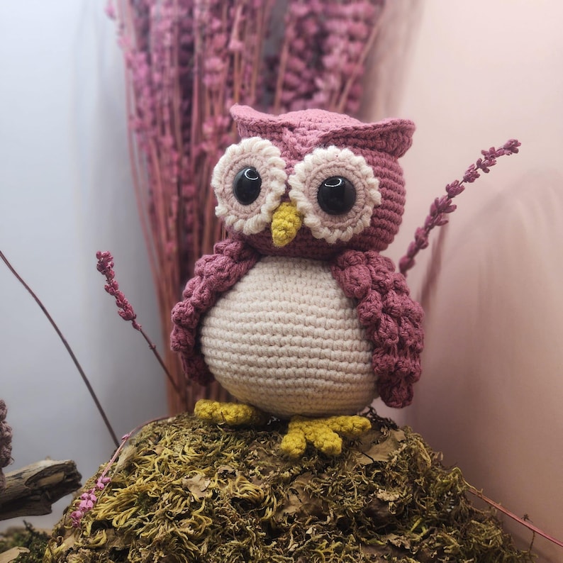 May include: A crocheted owl figurine featuring a dusty rose body, a cream-colored belly, and large black eyes surrounded by cream-colored circular patches. The owl has a small yellow beak and yellow feet, with textured wings and ear tufts.