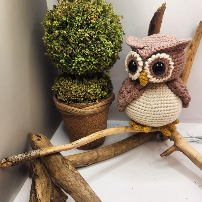 May include: A crocheted owl figurine with a dusty pink head and wings, a cream-colored belly, large black plastic eyes with white stitched borders, and a small yellow beak. The owl has yellow feet and is perched on a light brown wooden branch.