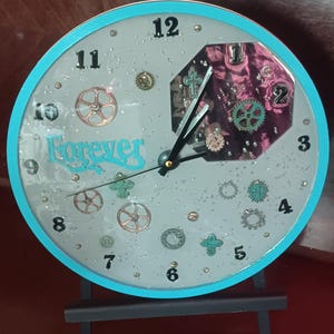 May include: A round clock with a light blue border and a gray face. The clock features black numerals, decorative gears, and the word "Forever" in turquoise script. The clock is mounted on a black stand.
