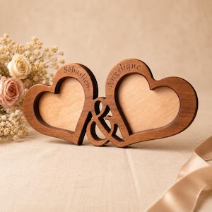May include: A double heart-shaped wooden photo frame featuring a central ampersand symbol. The left heart is engraved with the name Sébastien and the right heart is engraved with the name Angélique. The frame is crafted from dark brown wood.