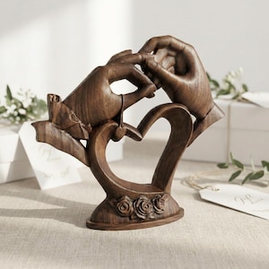 Può includere: Scultura in legno raffigurante due mani che si scambiano un anello, su una base a forma di cuore. La scultura è di colore marrone caldo, con dettagli intricati. La base presenta tre rose scolpite, a tema matrimonio o fidanzamento.