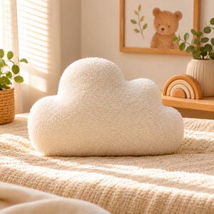 Könnte beinhalten: Ein flauschiges, wolkenförmiges Dekokissen in einem weichen Off-White-Ton. Das Kissen hat eine strukturierte, plüschige Oberfläche und liegt auf einer beigefarbenen Decke. Ein gerahmtes Bild eines Bären und ein Holzregenbogen sind ebenfalls zu sehen.