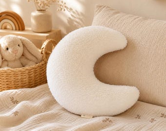 May include: A cream-colored, crescent moon-shaped pillow with a soft, textured surface. The pillow is placed on a cream-colored bedspread with a floral pattern. A wicker basket with a stuffed bunny and a wooden rainbow are in the background.