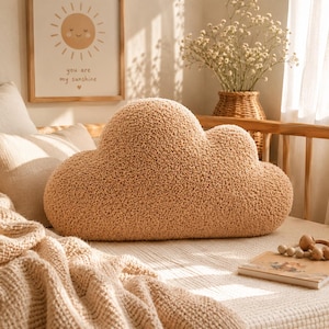 Puede incluir: Un cojín afelpado en forma de nube en un tono beige cálido descansa sobre una cama. El cojín tiene una superficie texturizada y difusa. Una imagen enmarcada con un sol sonriente y el texto "you are my sunshine" cuelga de la pared.
