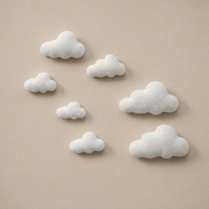 May include: Seven white, fluffy cloud-shaped decorations are arranged on a beige background. The clouds vary in size, with a textured, soft appearance. The arrangement suggests a whimsical, decorative theme, suitable for a nursery or children's room.