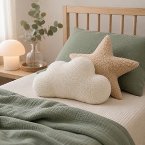 May include: A bedroom scene with a white cloud-shaped pillow and a tan star-shaped pillow. A sage green pillow and blanket are visible. A mushroom-shaped lamp and a vase with greenery are on a wooden bedside table.