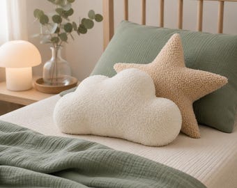 Könnte beinhalten: Ein Schlafzimmer mit einem weißen wolkenförmigen Kissen und einem tan-farbenen sternförmigen Kissen. Ein salbeigrünes Kissen und eine Decke sind zu sehen. Eine pilzförmige Lampe und eine Vase mit Grün stehen auf einem Nachttisch aus Holz.