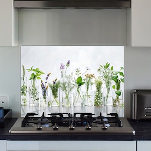 May include: A kitchen backsplash featuring a row of clear glass bottles, each holding different fresh herbs and flowers. The arrangement includes lavender, green herbs, and other colorful blooms. The backsplash is set above a black stovetop.