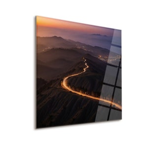 May include: A winding road with car headlights trails through a mountainous landscape at dusk. The sky is a gradient of orange, pink, and purple.