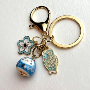 May include: A gold-colored keychain with a lobster clasp and a large ring. The keychain features three charms: a blue and white flower, a blue and white lucky cat, and a blue and white fish. The charms are detailed with gold accents.