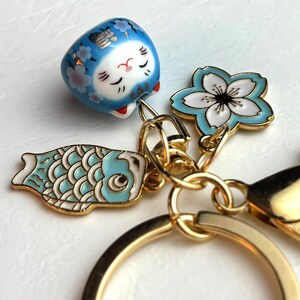 May include: A gold keychain with Japanese-themed charms. The charms include a blue Daruma doll, a light blue and white cherry blossom, and a light blue koi fish. The keychain has a gold ring and clasp.