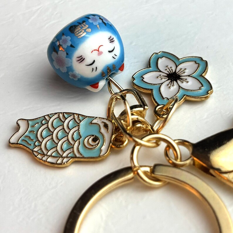 May include: A gold keychain with Japanese-themed charms. The charms include a blue Daruma doll, a light blue and white cherry blossom, and a light blue koi fish. The keychain has a gold ring and clasp.