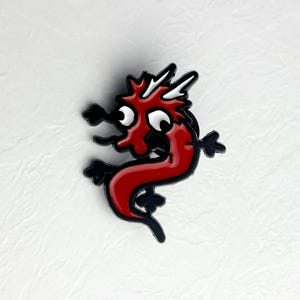 Op de afbeelding: Een rode drakenemaille pin met een zwarte omtrek. De draak heeft witte accenten, grote ogen en een grillig ontwerp. Deze pin is een leuk accessoire voor jassen, tassen of hoeden.
