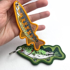 Puede incluir: Llaveros de peces bordados. Un llavero presenta una trucha marrón con detalles naranjas, amarillos y grises. El otro llavero es una lubina verde y blanca. Ambos llaveros tienen un anillo de metal para sujetar llaves.