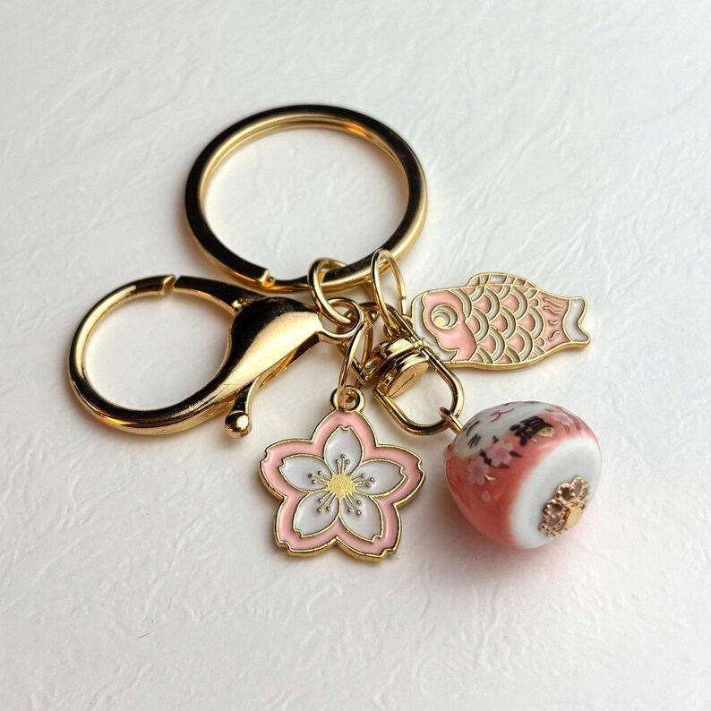 May include: Gold-toned keychain with a lobster clasp and key ring. The keychain features three charms: a pink and white cherry blossom, a pink fish, and a round, patterned bead with Japanese text. The charms are detailed with gold accents.