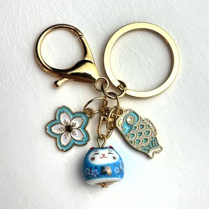 May include: Gold-tone keychain with a lobster clasp and a split ring. The keychain features three charms: a light blue and white flower, a blue and white fish, and a blue cat figurine with floral details. The cat figurine has a smiling face.