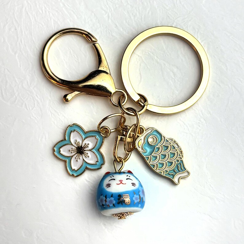 May include: Gold-tone keychain with a lobster clasp and a split ring. The keychain features three charms: a light blue and white flower, a blue and white fish, and a blue cat figurine with floral details. The cat figurine has a smiling face.