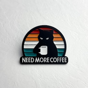 Può includere: Spilla con silhouette di gatto nero che tiene una tazza di caffè bianca, su sfondo color tramonto. Il testo "NEED MORE COFFEE" è visualizzato sotto. Un accessorio divertente per gli amanti del caffè.